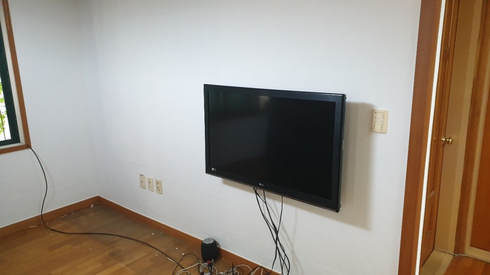 벽걸이 TV 아래로 전선이 내려온 흰 벽면과 작은 잔여물이 보이는 목재 바닥