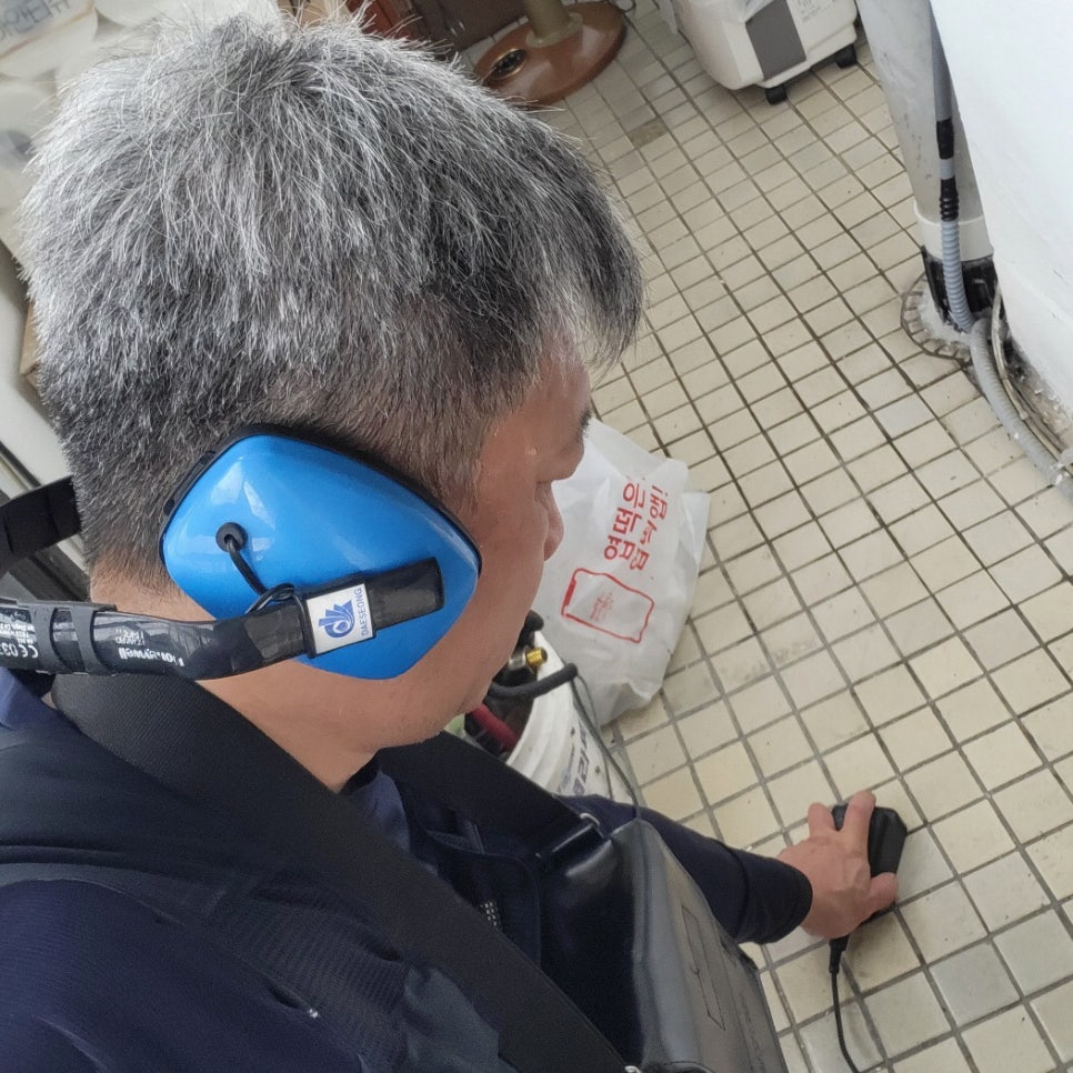 파란 헤드폰을 쓴 작업자가 타일 바닥에 유선 기기를 대고 있는 모습