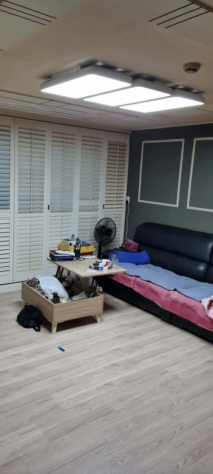 사각 조명 아래 흰 창 가림판과 검은 소파, 낮은 테이블이 있는 거실