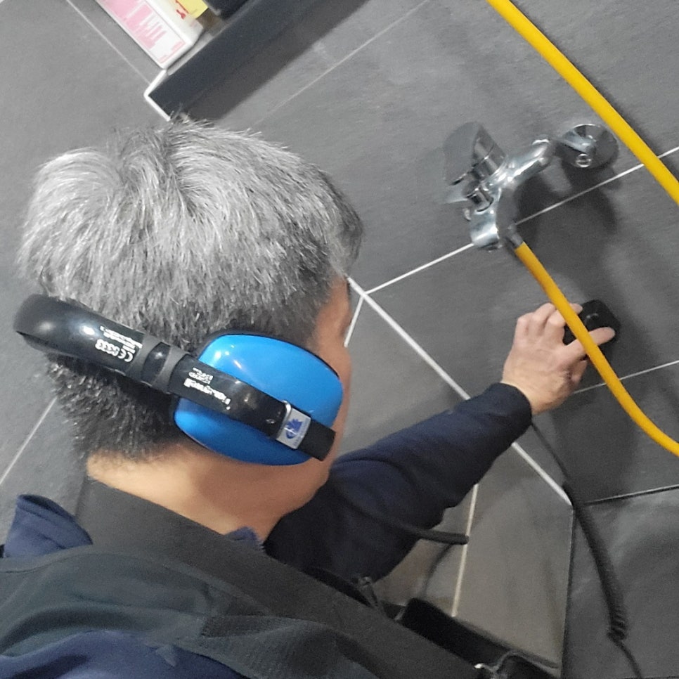 파란 헤드폰을 착용하고 수전 아래 벽타일에 검은 장치를 대는 작업자